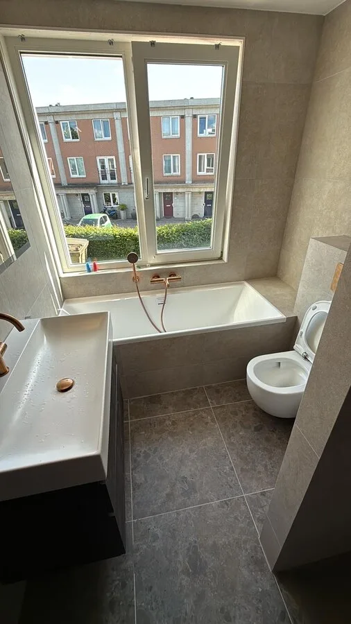 Moderne badkamer met ligbad, zwevend toilet en wastafel, beige tegels en koperen kranen.